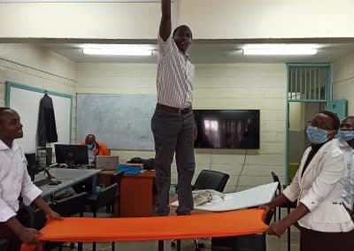 Un enseignant kényan debout sur une table tient un smartphoene en l'air. Devant lui ses collègues étendent un tapis orange