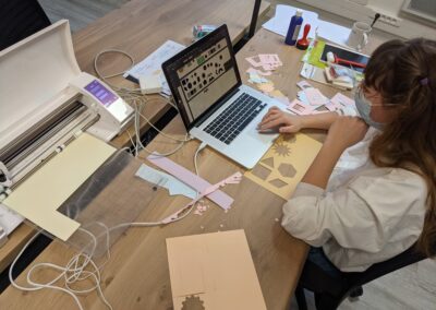 une étudiante travaille sur son ordinateur sur une table pleine de papiers découpés