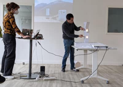 Une femme debout face à un ordinateur, un homme debout devant une table blanche manipule des objets lumineux